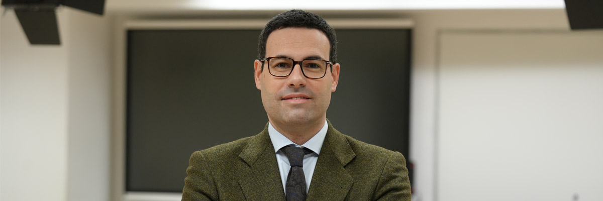 Retrato de Miguel Ortego, con gafas y chaqueta, de pie frente a una pizarra y pantalla en un entorno de aula