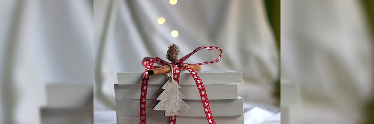Un regalo envuelto con una cinta roja y blanca decorada, adornado con un adorno de árbol de Navidad de madera y una piña, con luces desenfocadas en el fondo.