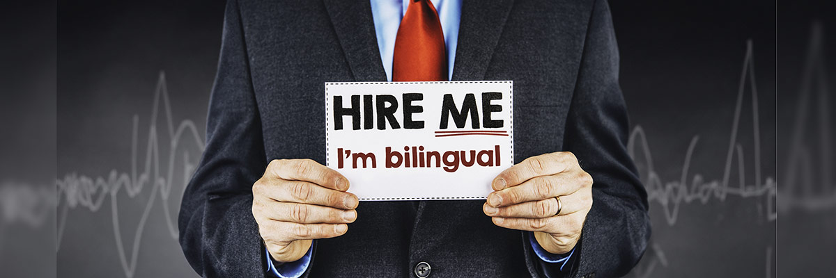 Una persona en traje sostiene un cartel que dice ‘CONTRÁTAME, soy bilingüe’ frente a un fondo de pizarra con gráficos.